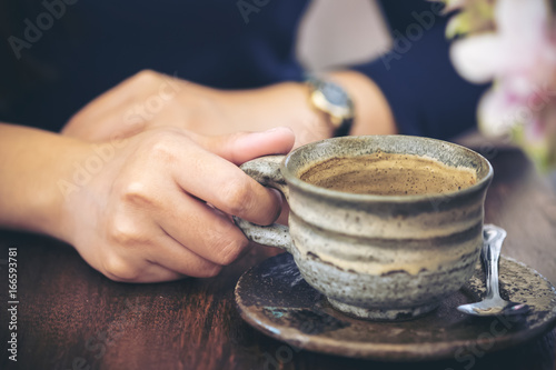 Closeup image of a woman's hand holding a cup of coffee on wooden table in vintage cafe