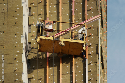 worker in yellow suspended cradle mounts environmental boards for insulation on a newly built high-rise building