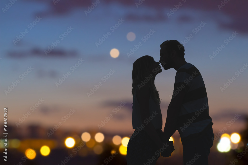 Silhouettes of a young couple kissing with city panorama in the background. - obrazy, fototapety, plakaty 