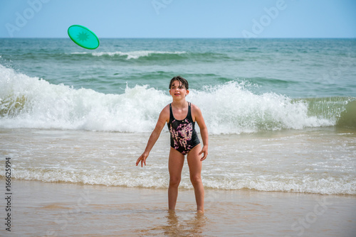 Cute youg girl in swimsuit standing in water with waves throwing a disc.