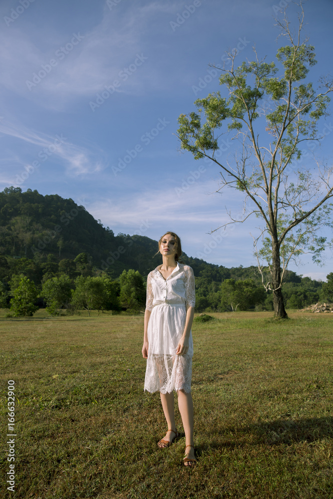 Pretty girl or cute woman, fashionable model standing with white strings in long, brunette hair, hairstyle in white dress on blue sky on mountain landscape. Fashion and beauty.
