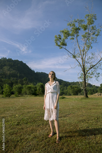 Pretty girl or cute woman, fashionable model standing with white strings in long, brunette hair, hairstyle in white dress on blue sky on mountain landscape. Fashion and beauty. © Bitmap
