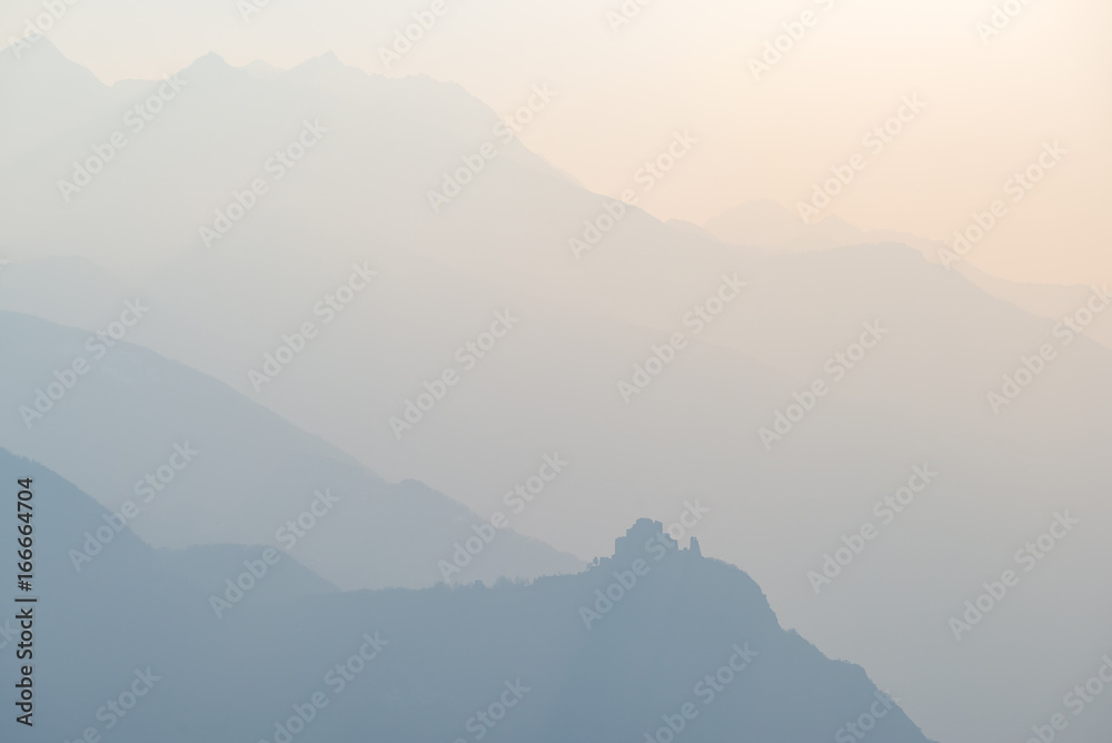 Fototapeta premium Blue toned distant mountain range of the Alps with St. Michel Abbey profile at the bottom. Torino Province, Piedmont, Italy. Telephoto view, soft light at sunset.