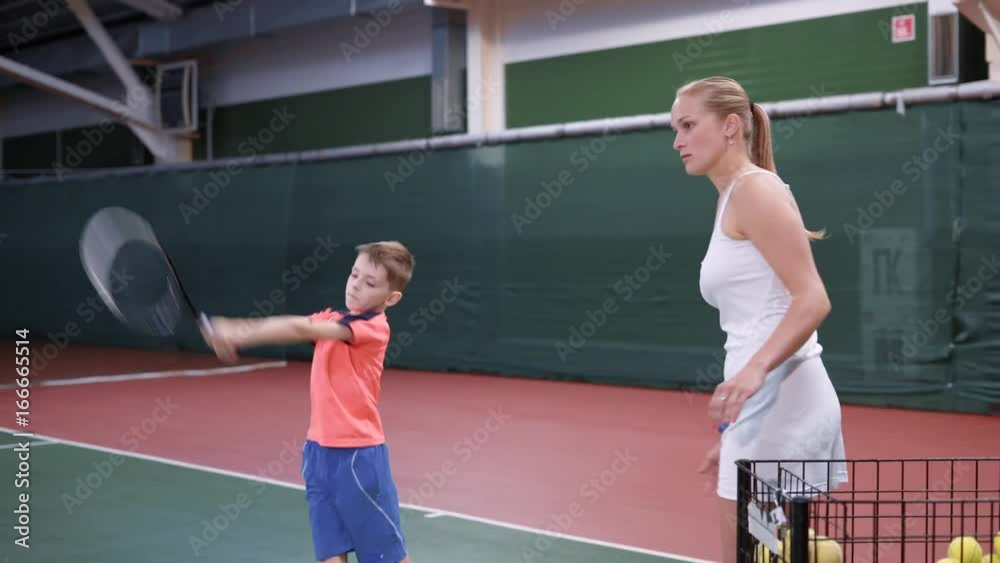Image of lesson at indoor court with professional equipment. Child learning process of correct using a racket. Boy playing tennis with female coach in white outfit giving him yellow balls. Image of lesson at indoor court with professional equipment. Child learning process of correct using a racket. Boy playing tennis with female coach in white outfit giving him yellow balls.