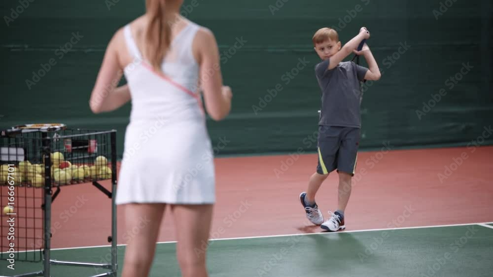 Female instructor teaching little boy playing tennis. Woman in white sport outfit standing in the court throwing yellow balls to child. Young sportsman learning to return tennis-ball with a racket. Female instructor teaching little boy playing tennis. Woman in white sport outfit standing in the court throwing yellow balls to child. Young sportsman learning to return tennis-ball with a racket.