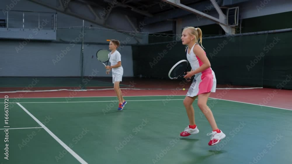 Two young athletes in recreation area playing sport game. Happy sister and brother having tennis lesson spending time at indoor court. Girl and boy serving and returning yellow balls with rackets. Two young athletes in recreation area playing sport game. Happy sister and brother having tennis lesson spending time at indoor court. Girl and boy serving and returning yellow balls with rackets.