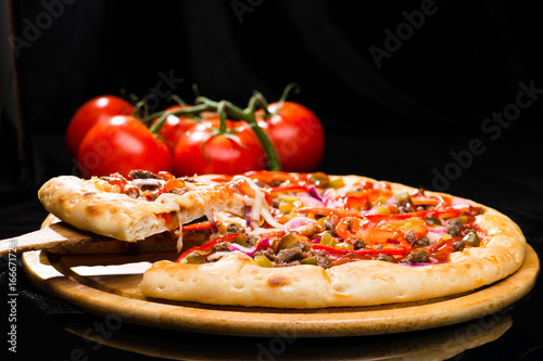 appetizing Italian pizza and sliced piece with meat, chili pepper, tomato sauce, pickled cucumbers and mustard on wooden board on dark background
