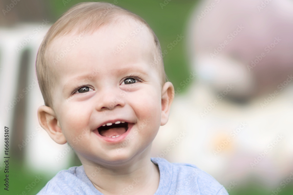 Close-up portrait of beautiful little baby boy laughing and looking to camera at summer day. Blurred background