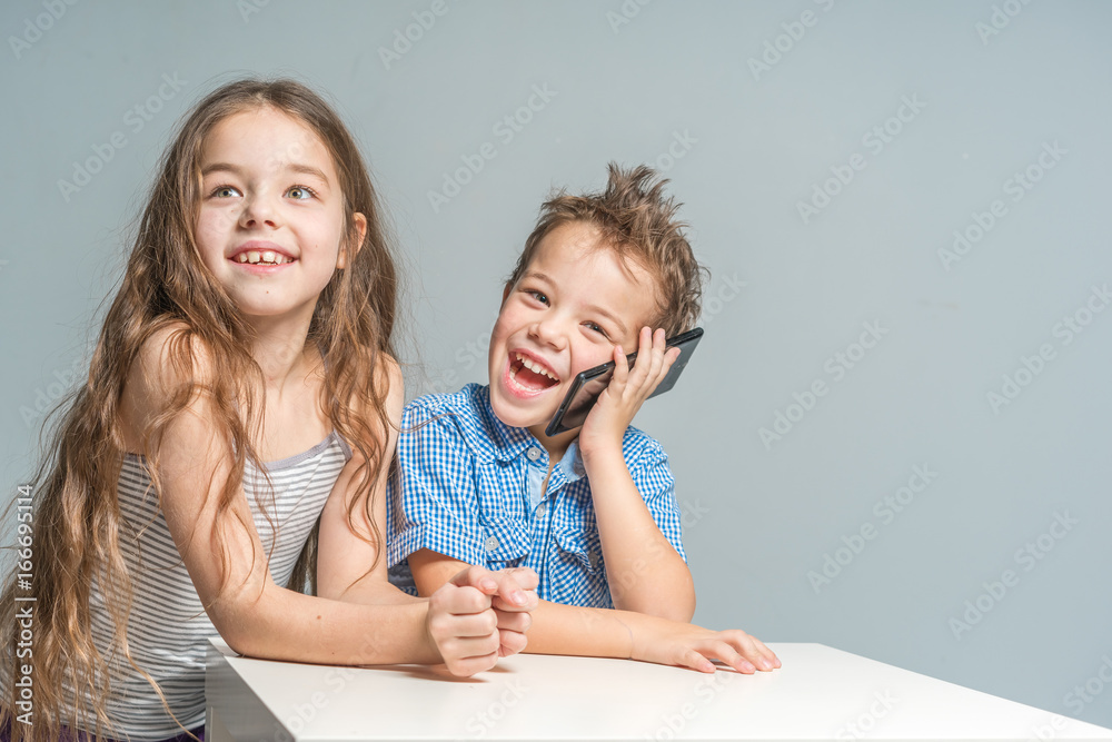 Smiling girl and boy with the phone sitting at the table on a gray background. Isolated Smiling girl and boy with the phone sitting at the table on a gray background. Isolated