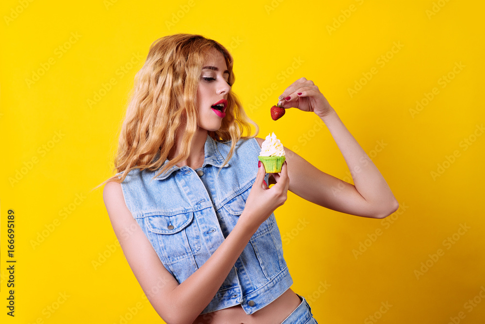 Fototapeta premium Attractive young woman trying to eat a cupcake with strawberry, over yellow background.