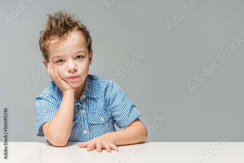 Cute thoughtful boy in a blue shirt at the table on a gray background. Isolated