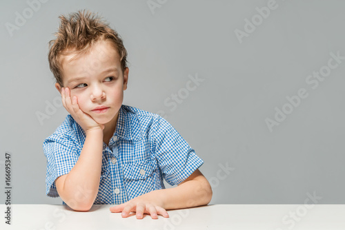 Cute thoughtful boy in a blue shirt at the table on a gray background. Isolated
