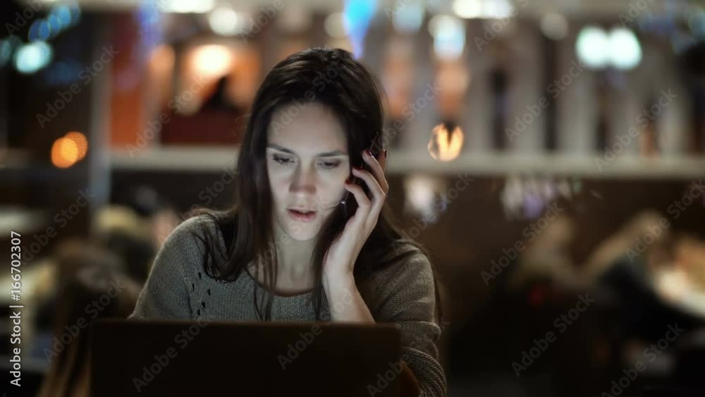 Young beautiful woman working on laptop in evening. Brunette female sitting in cafe and talking on smartphone. Young beautiful woman working on laptop in evening. Brunette female sitting in cafe and talking on smartphone.