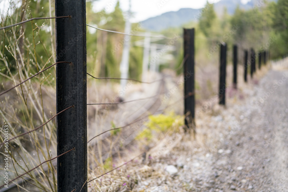 Rural fence dividing a country road from railway tracks with focus to the closest post and strands of wire over a blurred background in a receding perspective