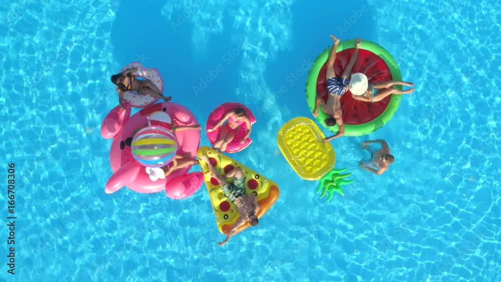 AERIAL TOP DOWN: Happy girls and guys playing with ball on floaties in pool. Cheerful smiling friends enjoying summer vacation on inflatable pineapple, pizza, flamingo, watermelon and doughnut floats