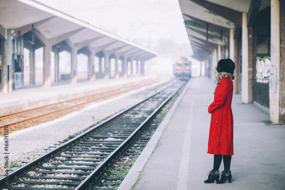Young beautiful woman curly hair in a red coat and russian fur hat on a train station Young beautiful woman curly hair in a red coat and russian fur hat on a train station
