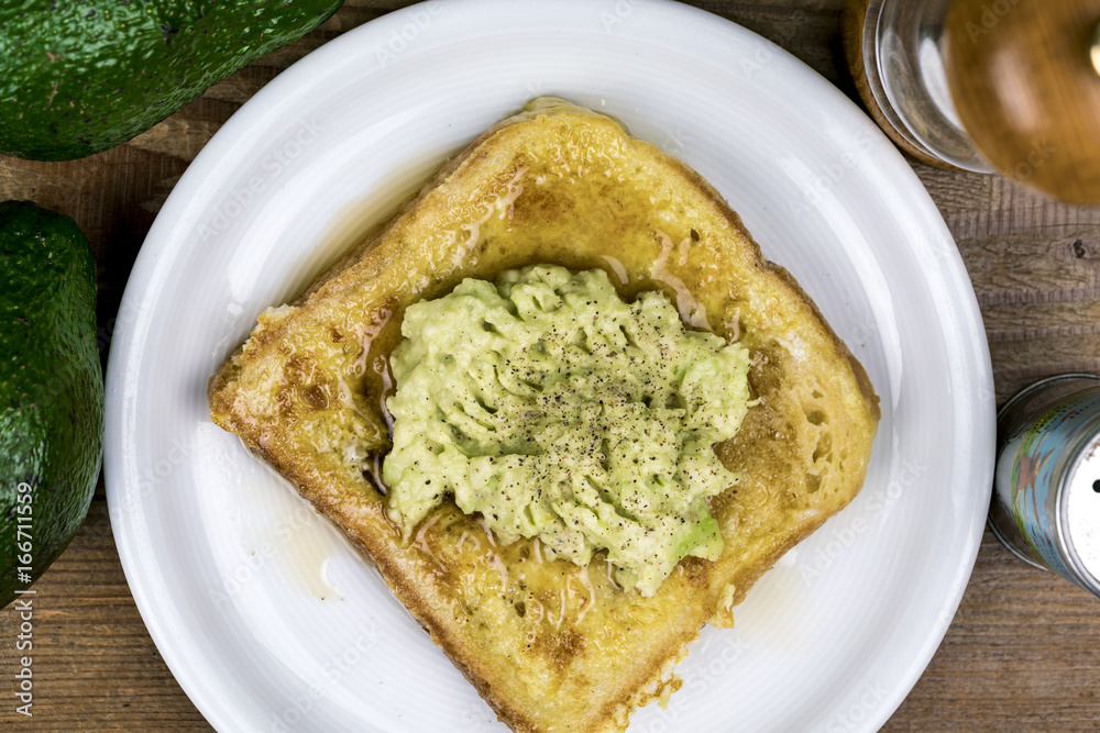 Mashed fresh avocado pear on a slice of toast with salt and pepper seasoning viewed from above on a white plate with ingredients Mashed fresh avocado pear on a slice of toast with salt and pepper seasoning viewed from above on a white plate with ingredients