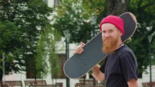 Young readhead hispter with beard and moustaches in jeans and T-shirt keeps skateboard on the soulder and walks the street looking to the camera. Outdoor. Cloudy. Medium. Stabilised. Portrait photo