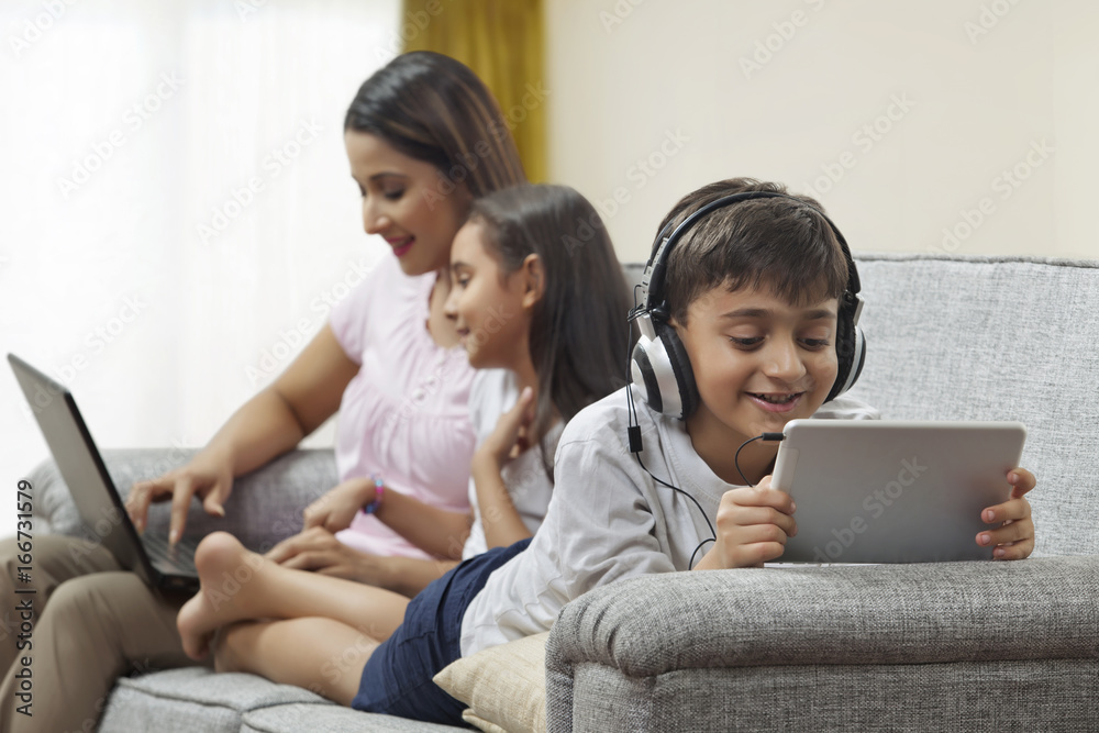 Focused boy using digital tablet headphones with mother and sister using laptop in background Focused boy using digital tablet headphones with mother and sister using laptop in background