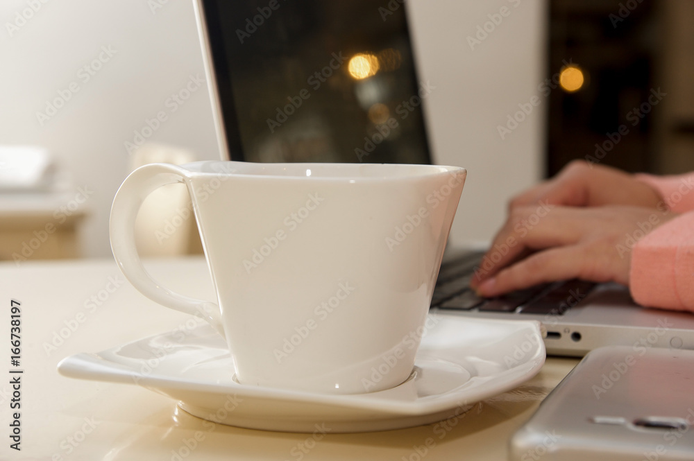 Close up of a white cup of coffe with a blurred businesswoman hand working in the background. Business concept Close up of a white cup of coffe with a blurred businesswoman hand working in the background. Business concept
