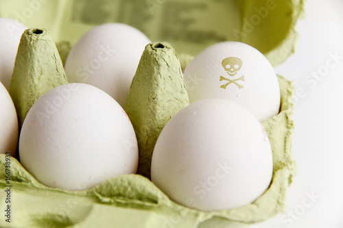 Eggs in a green paper package with one of the eggs painted with a poisonous risk symbol skull and bones. Image concept for food contamination.