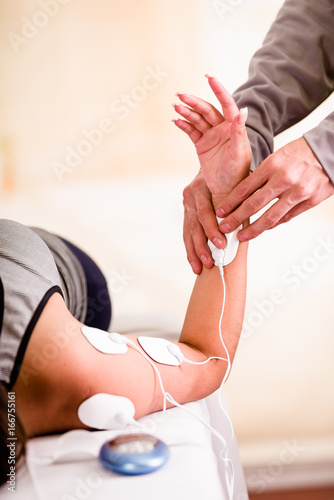 Close-up of a deportist holding with one hand the electric machine and putting the electrostimulator electrodes the arm of a female deportist