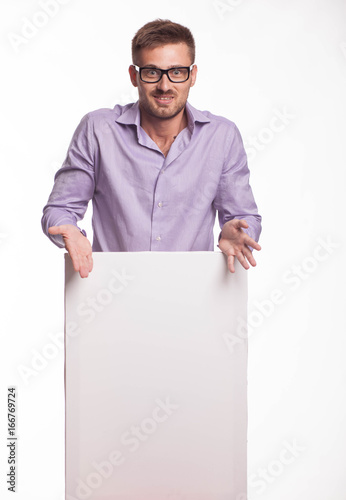 Young surprised man portrait of a confident businessman showing presentation, pointing paper placard gray background. Ideal for banners, registration forms, presentation, landings, presenting concept.