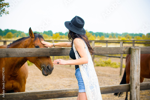 Woman in black hat with brunette curly hair and black dress and with horses on a farm, pets animals in village in a rancho. Horses are humans friends