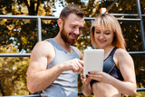 Young athletic woman and bearded man browsing the internet on tablet PC while doing fitness exercises in park.