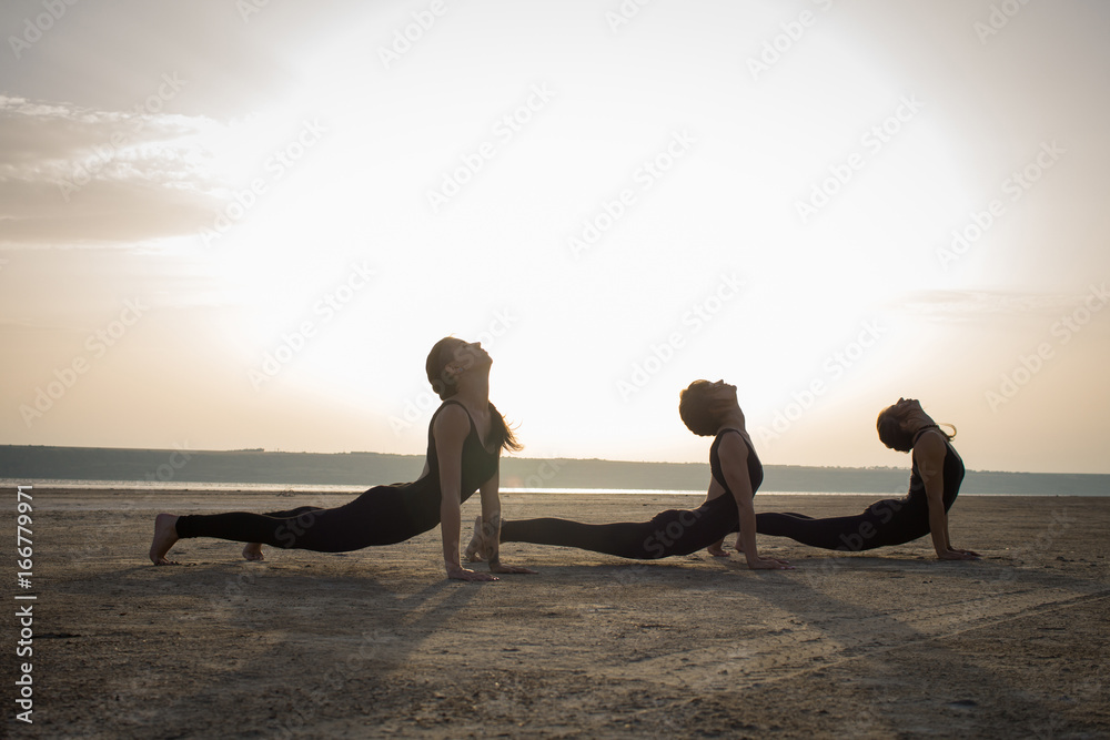 young women practicing yoga poses and asanas. Partner yoga, acrobatic yoga. Yoga class in black wear training in desert during sunset  