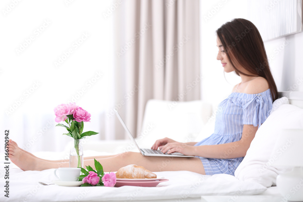 Tasty breakfast and young woman using laptop on bed in hotel room Tasty breakfast and young woman using laptop on bed in hotel room