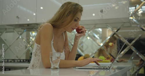 The close-up side portrait of the attractive blonde woman eating the apple while chatting via the laptop on the modern kitchen. photo