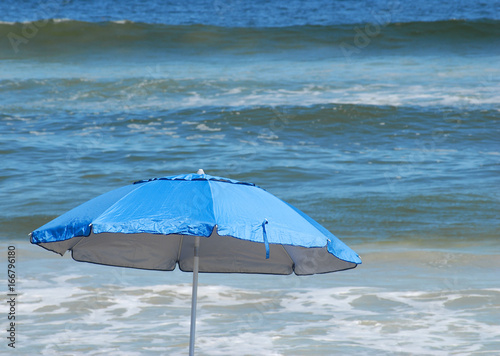 Selevtive focus is on a bright blue beach umbrella that is ruffled by the wind at the beach where waves can be seen rolling into shore © Perry Correll
