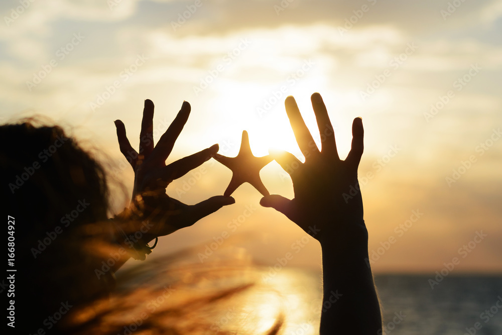 
Young, beautiful with long flying hair the girl holds the starfish in her hands on the background of the sunset at the sea shore. Romantic summer sea background 