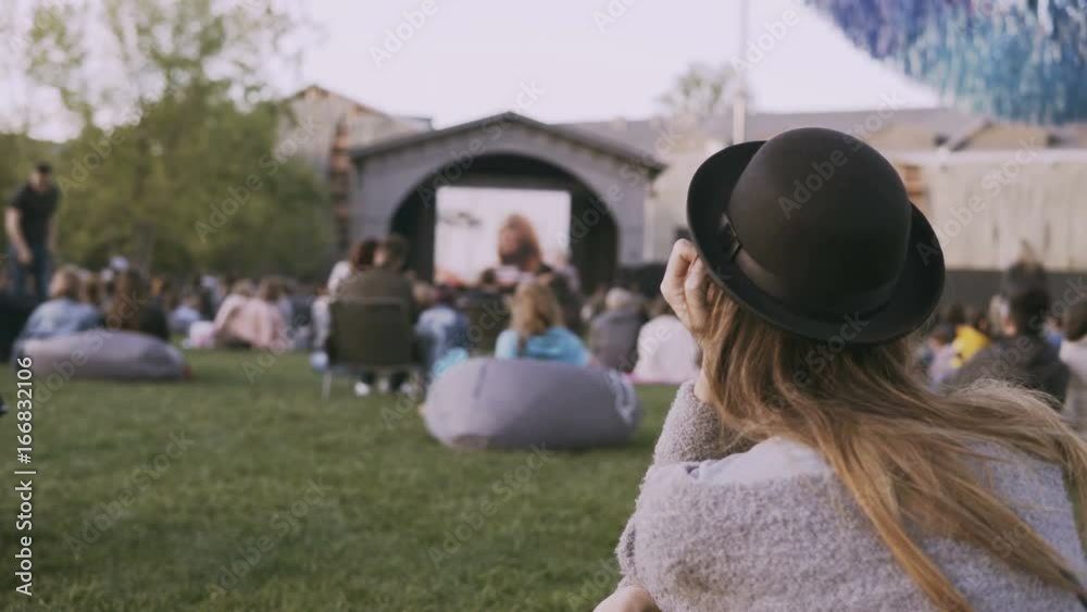 Back view of young stylish woman in hat sitting in bag-chair and looking movie in open-air cinema festival alone.