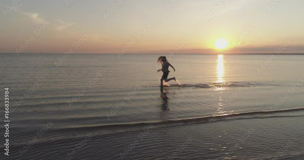 Aerial side fly over teen girl trying barefoot running in water on baltic sea beach in sunset time Aerial side fly over teen girl trying barefoot running in water on baltic sea beach in sunset time
