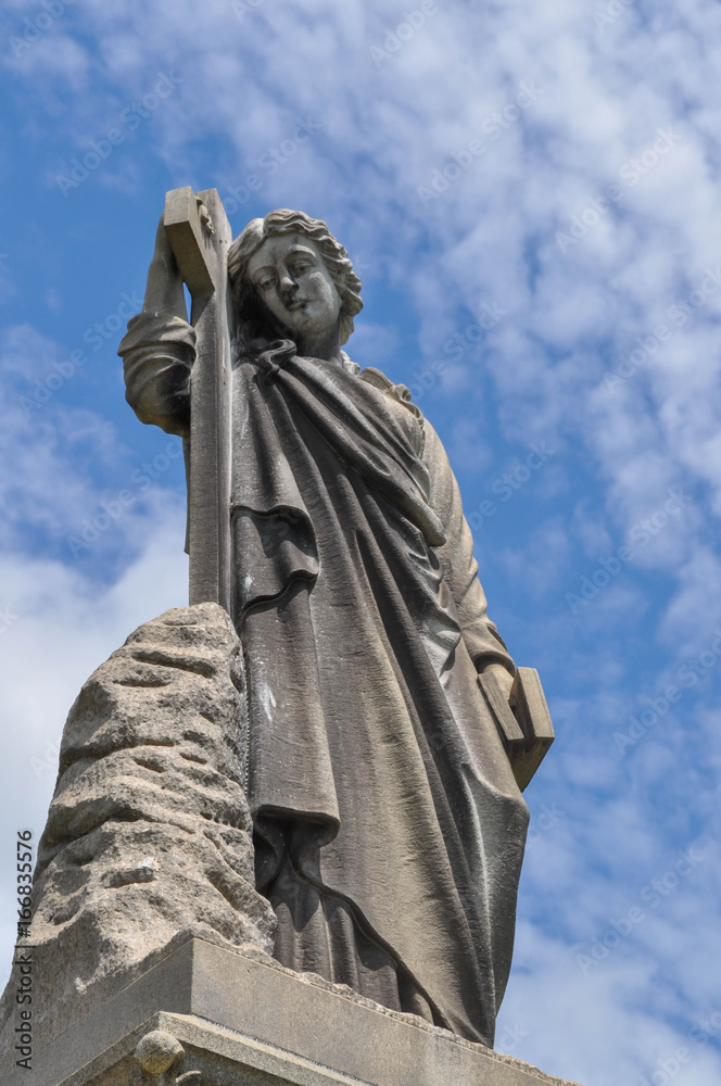 Fototapeta premium Cemetery statue of a person holding onto a book and a cross looking down upon a grave with a blue sky background