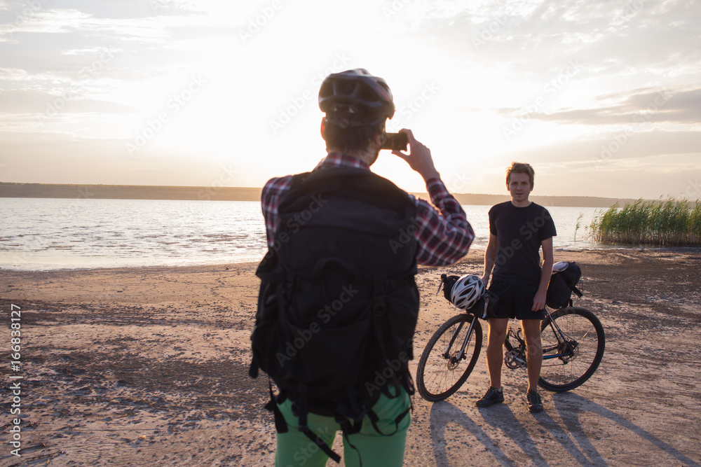 Fototapeta premium Tourist with backpack make photo of his friend with bicycle during sunrise in lake 