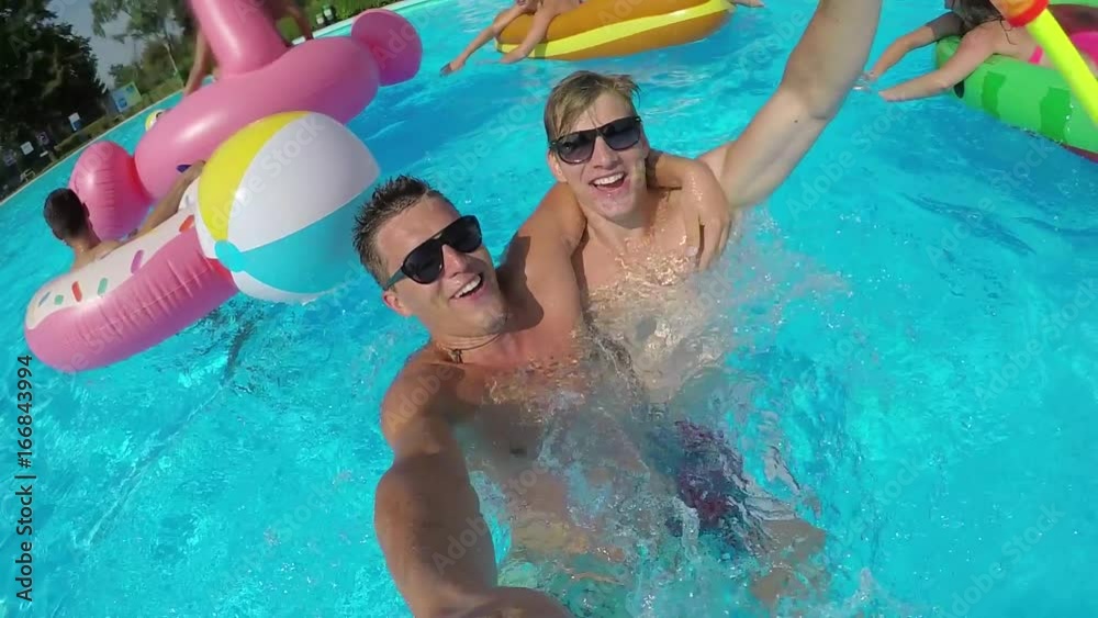 SLOW MOTION SELFIE Fun guys doing selfie video with smiling friends on colorful floaties in pool. Happy teenagers have water fight on summer vacation on inflatable pizza flamingo and watermelon floats