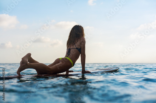 Happy girl in bikini have fun before surfing Surfer lie on surf board  look at sunset sky. People in water sport adventure camp  extreme activity on family summer beach vacation. Watersport background