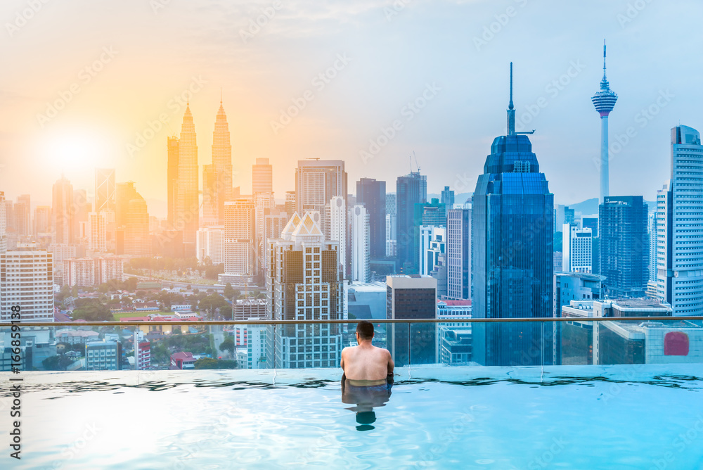 Naklejka premium Azjatycki biznesmen zrelaksować się w basenie na dachu za pięknym widokiem na miasto Kuala Lumpur w sunrise niebo, Malezja
