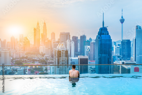 Canvas Print Asian businessman relax in swimming pool on roof top behind beautiful city view