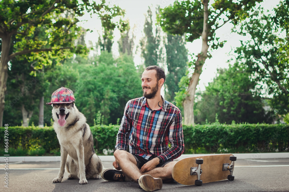 Young hipster man sitting on skateboard with funny siberian husky dog in cap 