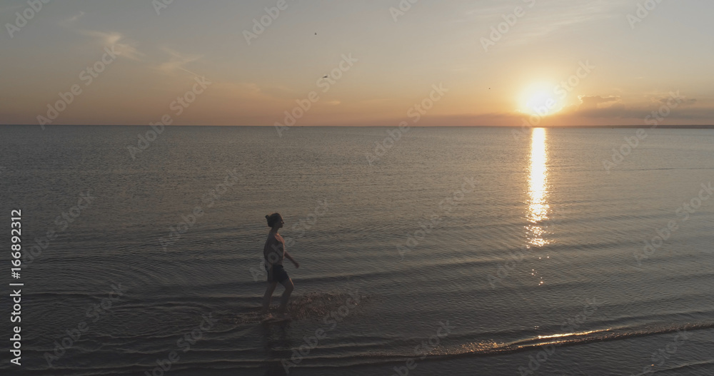 Aerial shot over teen girl barefoot walking in water on baltic sea beach in sunset time