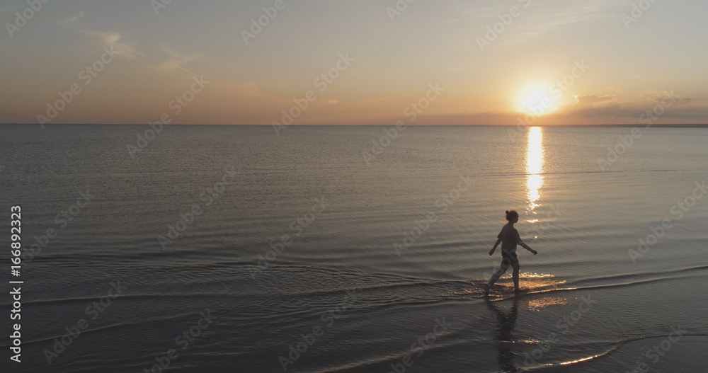 Aerial shot over teen girl barefoot walking in water on baltic sea beach in sunset time