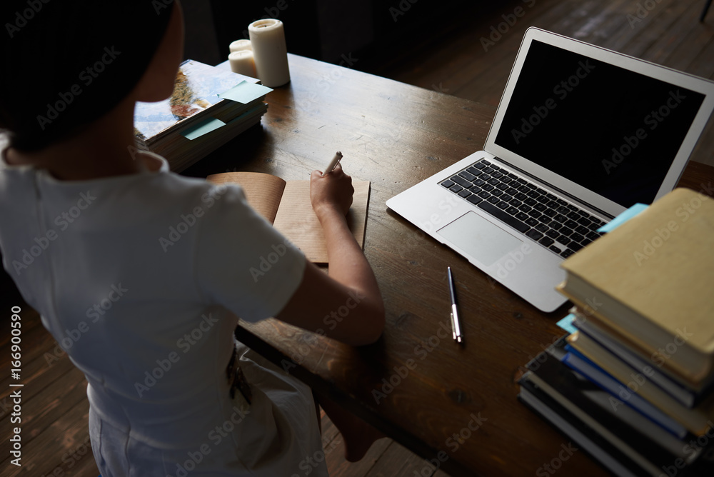 Unrecognizable female blogger making sketches in copybook, working at desk with piles of books and laptop with copy space black screen for your content. Woman handwriting in blank spiral notepad