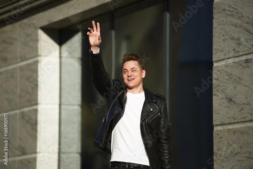 Fashionable smiling young European man wearing black leather jacket over white t-shirt standing outside modern buildin, waving hand while noticing his friend across street, trying to get attention