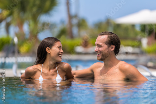 Happy swimming pool couple having fun laughing talking on luxury resort hotel travel holiday together on summer vacation. Asian woman, caucasian man in love on honeymoon.