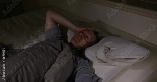 Mature man in bed cannot sleep at night time. He looks at alarm clock and tosses it in anger when it was time for morning wake up 
