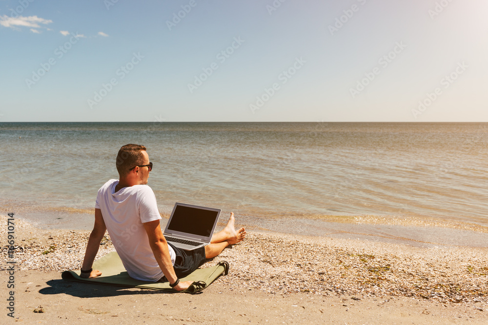 Young man in white T-shirt sits on the beach and working with laptop